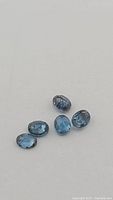 Five oval cut natural teal kyanite gemstones arranged closely on a light background.