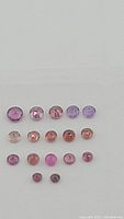 Clear close-up photo shows 15 round-cut natural garnet gemstones in a range of purple and pink shades, sizes between 2mm and 3mm, arranged in rows.