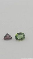 Close-up of two gemstones: one trillion cut deep red-purple-pink sapphire and one oval mint green tourmaline on white background.