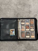 Open black binder showing 9 Magic: The Gathering cards in plastic sleeves and a sealed Magic deck pack inside the pocket