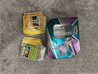 Lot contents showing two stacks of assorted Pokémon trading cards, some holo, and a purple/blue Pokémon tin box with Mew illustration and clear window on top.