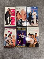 Top view of six VHS movies laid out on carpet floor, titles readable: Pretty Woman, Father of the Bride, Twins, 9 to 5, Second Chorus, and Three Men and a Baby.