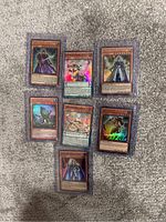 Seven Yu-Gi-Oh trading cards placed on a grey carpeted surface, showing different characters including holographic and non-holographic cards.