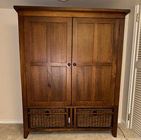 Full front view of the wooden armoire showing two paneled doors and two wicker baskets at the base.