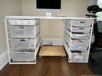 Full view of white Container Store Elfa desk showing dual drawer units with metal mesh drawers, wooden caster platform underneath, hardwood floor background.