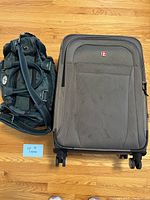 Photo showing both luggage pieces side by side on a hardwood floor: one large dark gray SwissGear rolling suitcase with visible wheels, and one soft carry-on bag in dark fabric next to it.