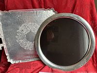 Two vintage trays: a round Kirk Stiff formica tray with pewter edge and a square forged aluminum floral tray, displayed on red fabric.