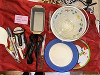 Overall photo showing various plates, bowls, kitchen tools, and utensils arranged on red cloth