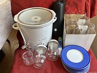 Top view photo of white Proctor Silex crockpot with glass lid, six stemless wine glasses in box, stack of blue-rimmed plates, and four cocktail jars in front. Red fabric background.
