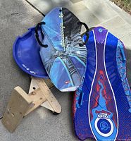 Four winter snow riding items including two snowboards with handles, a round blue plastic sled, and a handmade wooden mono-ski with seat and single ski base.
