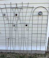 Three metal garden trellises stood against a house siding, showing decorative elements including metal leaves and a circular butterfly stained glass insert.