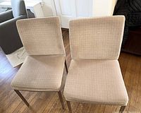 Two beige fabric dining chairs with wooden legs, placed on wood floor near window and door.