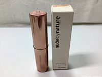 Nude By Nature Touch Of Glow Highlight Stick in rose gold tube next to its branded box with text and weight on a fabric background.