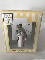 Front view of the new old stock boxed holiday ornament featuring a gray and white cat peeking out of a red and white striped container with green gift boxes on top.