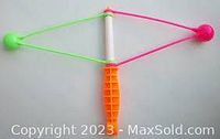 Top view photo of the vintage 1980s Klik Klak balls noise maker toy showing two neon plastic balls (green and pink) connected by elastic strings to a white plastic rod with an orange handle below.