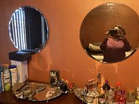 Two round wall mirrors are shown mounted on an orange wall above a dresser with various personal items. One large mirror reflects a person taking the photo, the other mirror is smaller with a visible silver frame.
