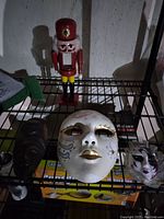 Porcelain masks and wooden nutcracker doll on metal shelf. Large porcelain mask is white with gold and colored paint detail, smaller mask is purple and white, nutcracker doll is classic style with red hat.