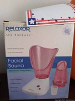 Front side of the Relaxor Spa Therapy Facial Sauna retail box showing the pink facial sauna device, inhaler attachment, measuring cup, branding and usage information.