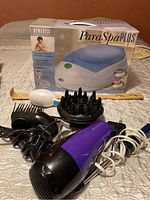 Photo of ParaSpa Plus paraffin bath with box, purple and black hair dryer, diffuser attachment, three black hair styling attachments and wooden spatula on table.