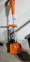 Electric snow shovel in orange and white with visible power cord on concrete floor against wall.