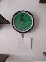 Photo of the round wall clock with green background and black frame hung on a white wall with wooden shelves nearby.