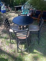 Outdoor photo of round black bar top table with four wooden slatted back bar chairs surrounding it on grass ground, showing wear and rust spots.