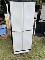Closed view of gray plastic HDX garage cabinet showing four vertical doors with black trim and base, standing on grass outdoors.