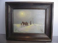 Framed original oil painting on board showing a winter scene with snow, a horse-drawn sleigh with two passengers, a standing figure, and crosses in the background under a pale glowing sun or moon.