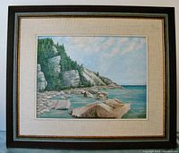 Framed oil painting showing rocky shoreline with pine trees and water under a soft cloudy sky.