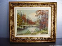 Framed oil painting showing the full autumn landscape artwork with trees and river, framed with an ornate gold and white frame.