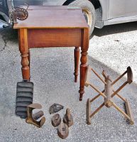 Antique wooden school desk with slanted lift top, detached from base. Various cast iron items including a wool winder, cornbread pan, sad irons, trivet, apple peeler, and other iron tools placed around the desk.