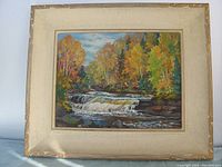 Oil painting showing waterfall with autumn trees in the background inside a cream and gold colored frame, showing minor signs of wear on the frame edges.
