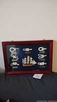 Overall view of the framed boat knot picture with seven rope knots arranged around a miniature ship, all mounted on dark blue fabric inside a reddish wood frame.