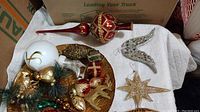 Top view of a shallow white bowl holding assorted gold, red, and green glass Christmas ornaments including finials, stars, and baubles, with some clear glass bird ornaments placed nearby on a white tablecloth.