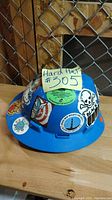 Blue hard hat decorated with various stickers placed on a wooden surface next to a chain link fence.