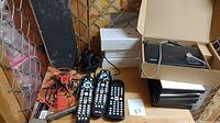 Mini skateboard deck, multiple remote controls, power adapters, empty iPhone boxes and black DVD boxes arranged on a shelf against a chain link fence
