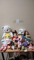 Photo of lot of stuffed animals including white bear, yellow character, lamb, dolls and various plush animals on table against wall.