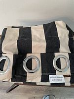 Two folded drapery panels with large black and sand stripes showing metal grommets and Ballard label