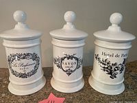 Photo showing all three white ceramic apothecary jars with lids, black decorative French text and designs, placed side by side.
