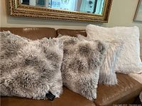 Four fluffy decorative pillows on a brown leather couch including three neutral beige long shaggy fur pillows and one white fluffy pillow.