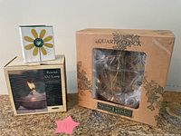 Three boxed items on a counter: Quartz Clock from Stone Garden in original box, pewter oil lamp boxed, and small garden book with flower on cover.