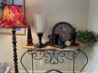 Wide view of entire home decor lot on a metal and stone table including the black floor lamp with floral shade, candle holder, horse head bookends, wall clock, finials, and artificial plant.