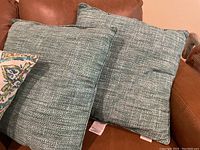 Two light green-blue teal pillows on brown leather couch, showing fabric texture and size.