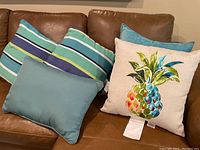 Five outdoor decorative pillows arranged on a brown leather couch: one pineapple print pillow, two aqua solid pillows, two striped pillows
