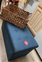 Blue rectangular upholstered storage bench with button tufted top, large rectangular woven basket sitting on top, kitchen setting with light wood floor.