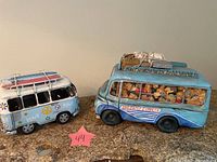 Side by side view of two colorful VW-style van figurines on granite surface against beige wall. Left is a blue peace van with surfboards on roof, right is the blue Cote d'Azur Express bus with caricature figures inside windows.