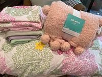 Stacked bedding including Tommy Hilfiger comforter, pillowcases, and shams with a folded pink Pillow Fort throw on top.