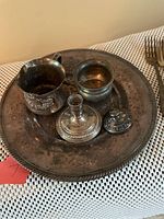 Shows the vintage silver plated round tray with decorative edges and four small silver plated items arranged on it: a small creamer, a small bowl, a candleholder with lid, and a lidded container. Tarnished and aged appearance visible.