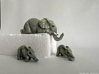 Three miniature elephant family figurines set on and around white foam packaging, showing the large elephant and two smaller baby elephants with textured, wrinkled skin and grayish-green paint.