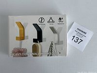 White package showing three wall hooks in yellow, black and white with use example.